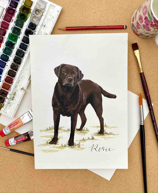 A detailed hand painted portrait of a brown Labrador dog, surrounded by paint palette, water jug and paint brushes.