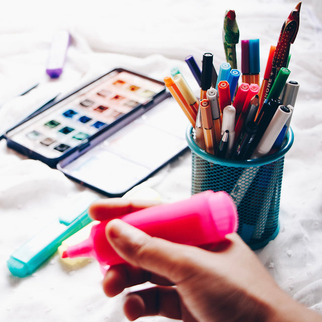 A hand holding a pink highlighter pen alongside a pot of pens and pencils, laid beside a watercolour paint palette.